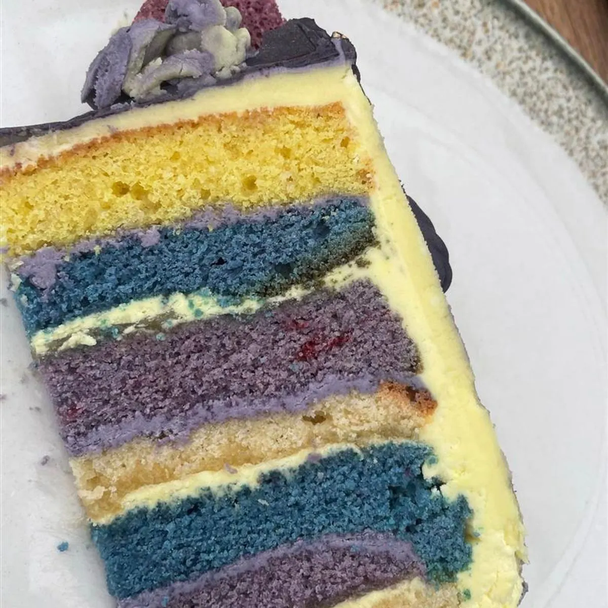 A slice of rainbow layer cake showing yellow, blue and purple sponge layers