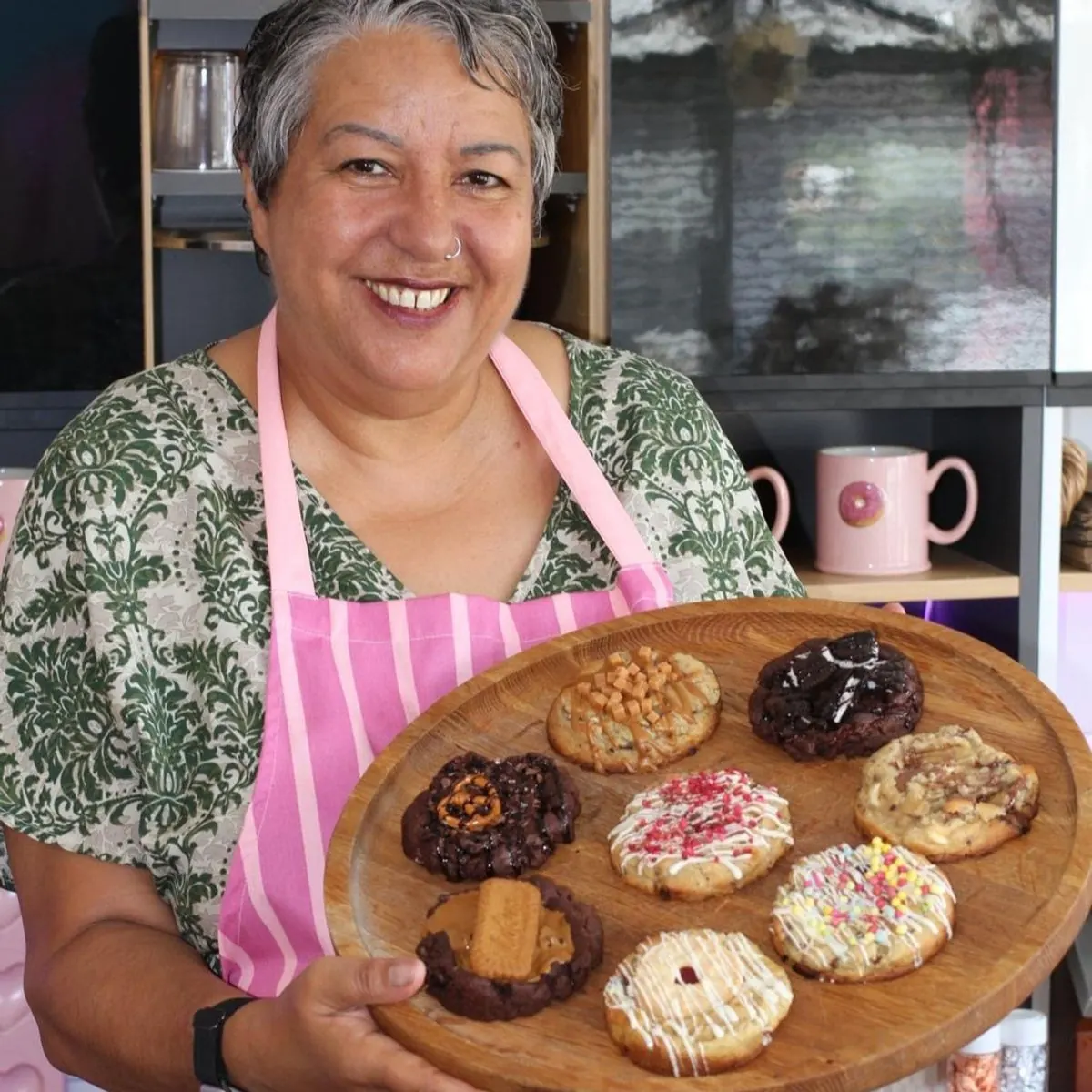 Tracy in a pink striped apron, smiling and holding a board of decorated cookies
